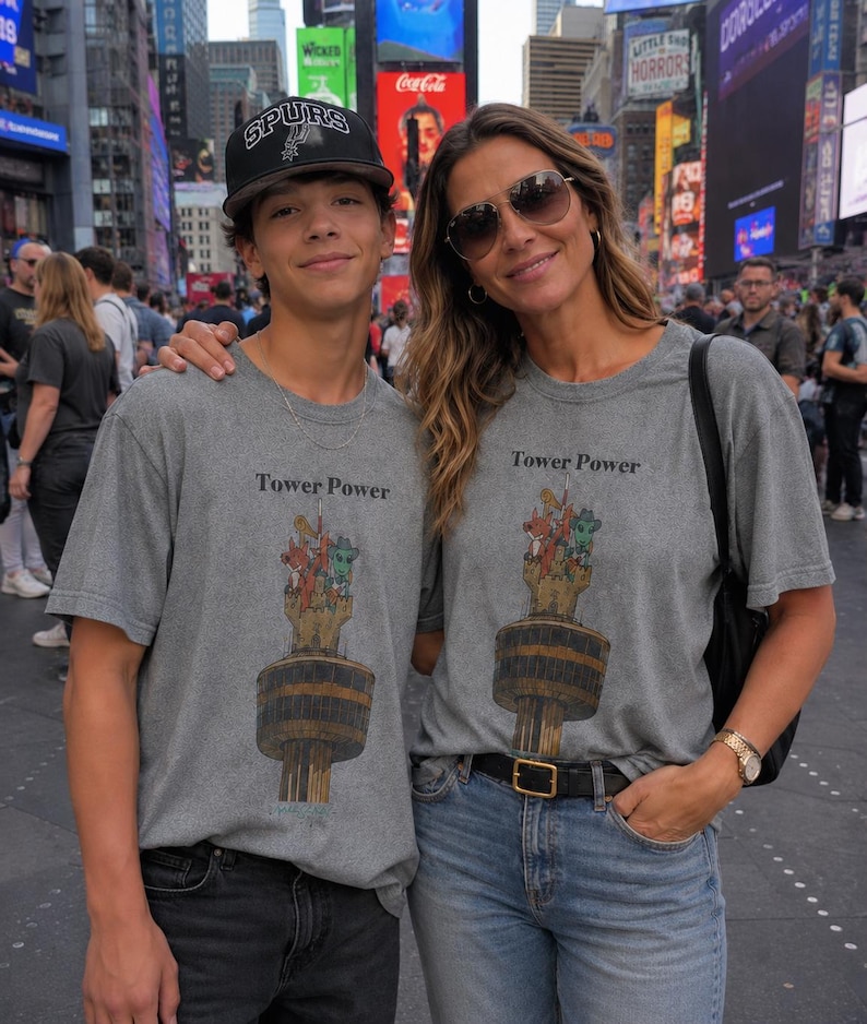 May include: Two grey crew-neck t-shirts featuring a graphic print of a tall tower with cartoon characters at the top and the text Tower Power. The shirts are worn with black jeans, a black baseball cap, blue jeans, and a black leather belt.