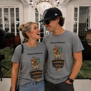 May include: A grey crew neck t-shirt featuring a graphic print of a tall tower with a fox and an alien character at the top. The text Tower Power is printed in black above the illustration. The design is centered on the chest of the garment.