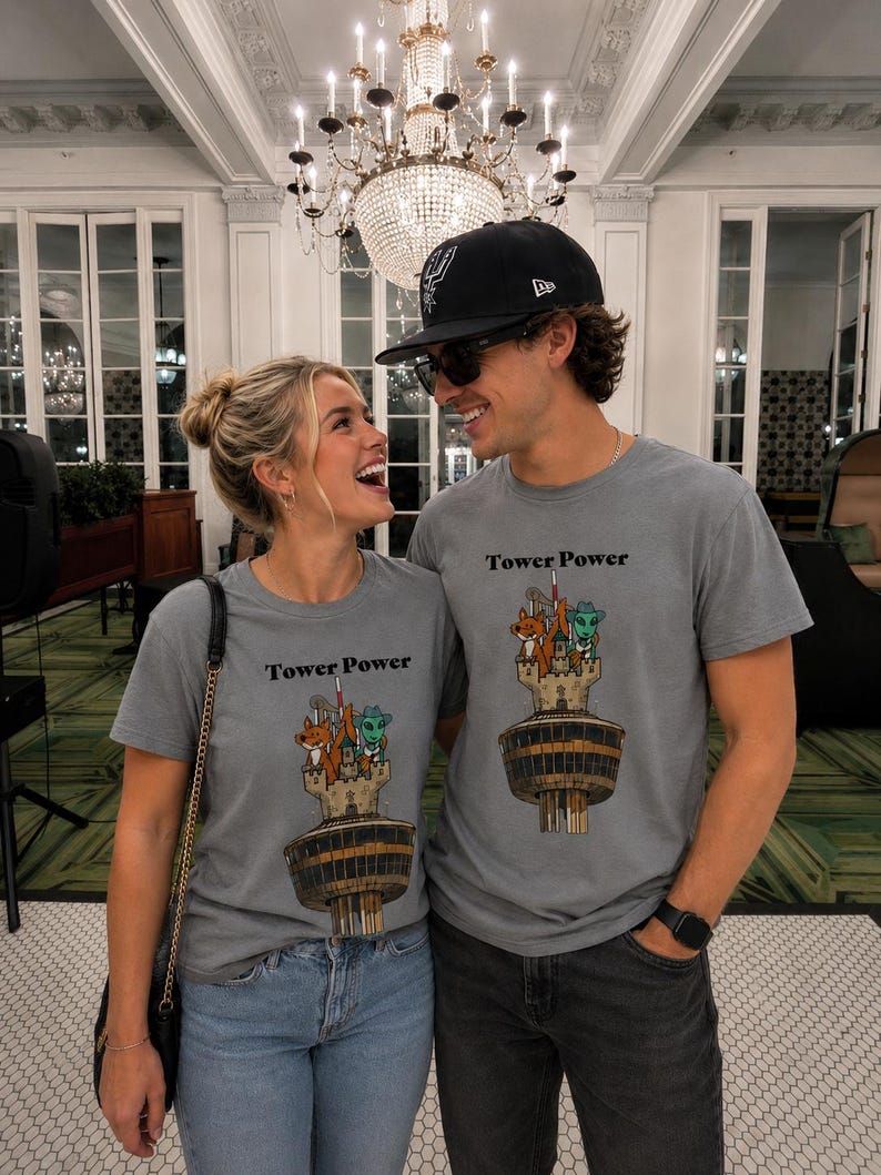 May include: A grey crew neck t-shirt featuring a graphic print of a tall tower with a fox and an alien character at the top. The text Tower Power is printed in black above the illustration. The design is centered on the chest of the garment.
