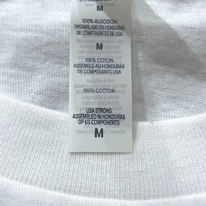 May include: A white fabric garment featuring a vertical interior tag with black text. The tag displays care instructions, the size M, and states 100% cotton, assembled in Honduras of US components. The fabric has a visible ribbed texture.