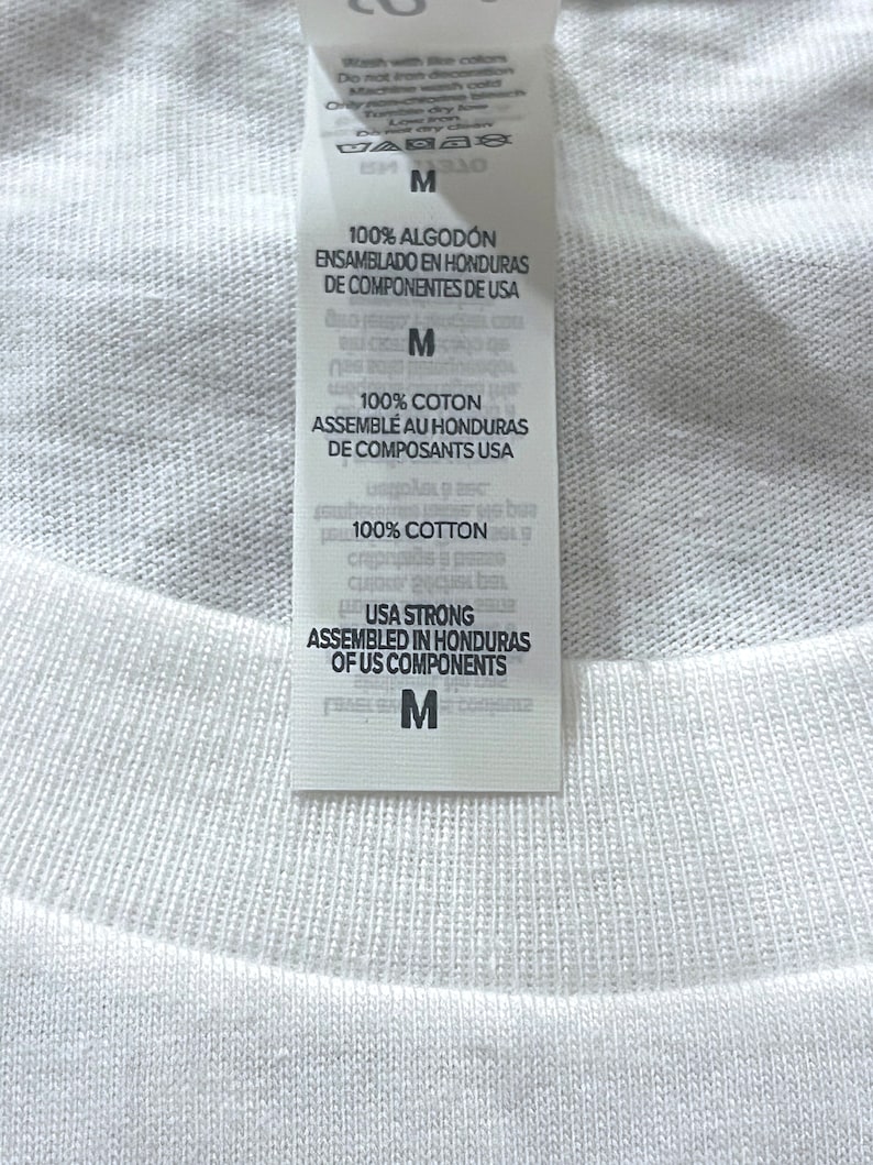 May include: A white fabric garment featuring a vertical interior tag with black text. The tag displays care instructions, the size M, and states 100% cotton, assembled in Honduras of US components. The fabric has a visible ribbed texture.