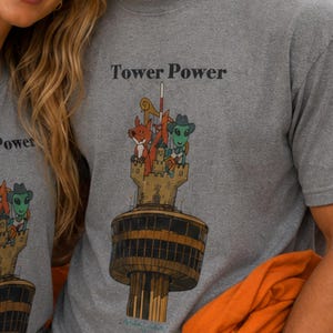 May include: A grey t-shirt with a subtle repeating pattern and a graphic print featuring the text Tower Power. The illustration depicts a tall tower topped with a castle structure, a fox, and a green alien wearing a cowboy hat and scarf.