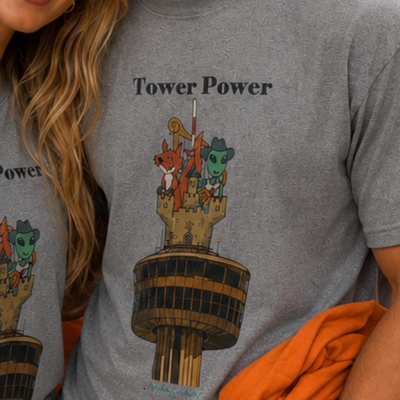 May include: A grey t-shirt with a subtle repeating pattern and a graphic print featuring the text Tower Power. The illustration depicts a tall tower topped with a castle structure, a fox, and a green alien wearing a cowboy hat and scarf.