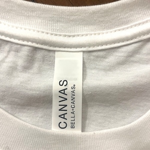 May include: A white t-shirt featuring a vertical fabric neck label. The label is white with black text that reads CANVAS BELLA+CANVAS. The shirt has a ribbed crew neckline and is constructed from a plain white textile material with visible stitching.