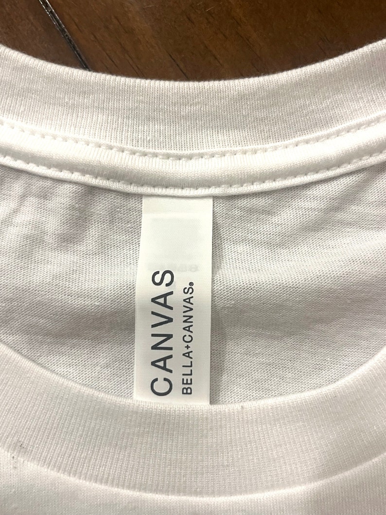 May include: A white t-shirt featuring a vertical fabric neck label. The label is white with black text that reads CANVAS BELLA+CANVAS. The shirt has a ribbed crew neckline and is constructed from a plain white textile material with visible stitching.