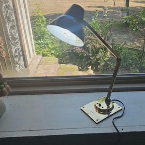 May include: A desk lamp with a black conical shade and a white interior. The lamp features a metallic gold-toned adjustable arm and a square base with a black power cord. The light is turned on, illuminating the white interior of the shade.