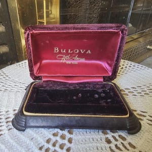 May include: Open vintage Bulova jewelry box with a deep burgundy velvet interior and exterior. The box features the Bulova logo in gold lettering on the inside lid. The box is rectangular with a hinged lid and a gold trim.