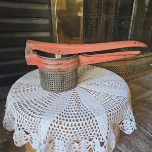 May include: An antique potato ricer with a red painted handle and a perforated metal basket. The kitchen tool is resting on a white lace doily.