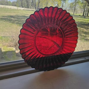 May include: A vibrant red glass plate with a scalloped edge. The plate has a ribbed design radiating from the center. The plate is positioned on a windowsill, with a natural outdoor background visible.