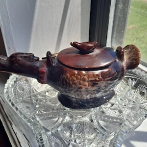 May include: A brown ceramic oil lamp with a bird-shaped lid. The lamp is decorated with a floral pattern and has a spout on the side. The lamp is sitting on a clear glass plate.