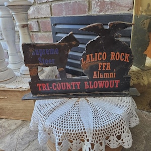May include: A vintage wooden sign with a cow silhouette and an eagle emblem. The sign features text in blue and red, including "Supreme Steer," "Calico Rock FFA Alumni," and "Tri-County Blowout."