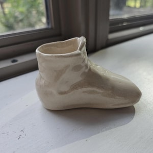 May include: A small, cream-colored ceramic figurine shaped like a simple boot or shoe. The object has a smooth, glossy finish with a hollow opening at the top, featuring a rounded toe and a slightly raised ankle section with a plain, unadorned surface.
