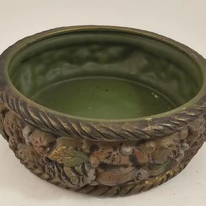 May include: A round, antique-style planter with a green interior and a decorative exterior. The planter's rim is a dark brown with a rope-like texture. The exterior features a relief design of fruits and foliage in shades of brown, green, and gold.