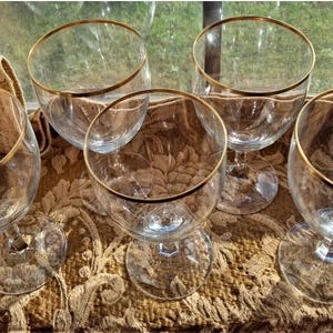 May include: Set of six clear glass wine glasses with a gold rim. The glasses are arranged on a brown patterned surface.