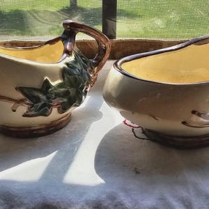 May include: Two vintage cream and sugar serving dishes. The cream pitcher has a handle and a spout. Both pieces are beige with brown trim and green leaf accents. The interior of the dishes is yellow.
