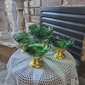 May include: Four green glass dessert bowls with scalloped edges and gold-colored pedestal bases are arranged on a white lace tablecloth. The bowls have a vibrant, emerald green color. The background includes a brick wall and decorative white candlesticks.