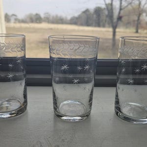 May include: Three clear drinking glasses with etched star and leaf patterns. The glasses are cylindrical with a slightly flared top. The etched designs are white against the clear glass. The glasses are empty and sit on a white surface.