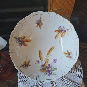 May include: White ceramic serving platter with a scalloped edge and gold trim. The platter is decorated with wheat and purple floral designs. The platter is sitting on a white lace doily.