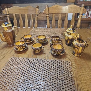 May include: A ceramic tea set featuring a tall teapot, a lidded sugar bowl, a creamer, and four teacups with matching saucers. Each piece displays a brown and yellow drip glaze finish with dark brown accents around the rims and handles.