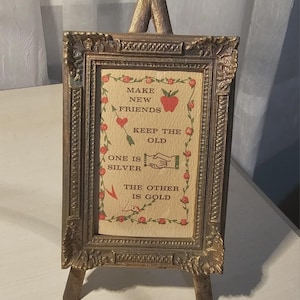 May include: A vintage-style framed print on a small easel. The frame is ornate with a gold finish. The print features the text "Make New Friends, Keep the Old, One is Silver, The Other is Gold" with decorative hearts, arrows, and an apple.