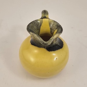 May include: A small, round yellow pitcher with a black and gray rim and handle. The pitcher has a bulbous body and a spout for pouring. The handle is a small, curved shape. The pitcher is likely ceramic or pottery.