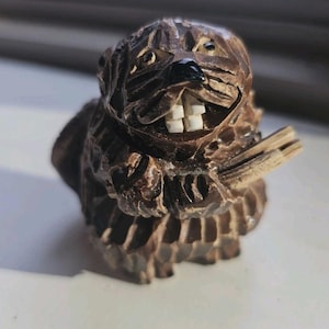 May include: A detailed, hand-carved wooden beaver figurine. The beaver is brown with white teeth and a black nose. It is holding a small piece of wood. The carving has visible texture and detail, showcasing the craftsmanship of the piece.