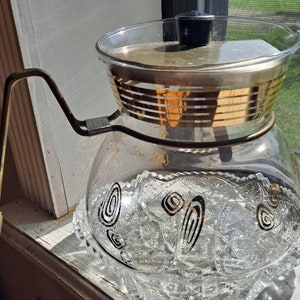 May include: A vintage glass coffee pot with a gold band around the top and a black and white swirl design on the bottom. The pot has a glass lid and a metal handle.