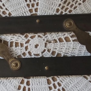 May include: Two dark wooden antique hinges with brass hardware. Each hinge has a rectangular wooden base and a decorative brass arm. The brass hardware includes circular accents and connecting pieces. The hinges are positioned on a white crocheted surface.