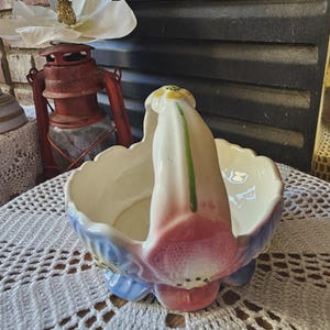 May include: A vintage ceramic basket with a handle, featuring a floral design in pastel colors. The basket has a scalloped rim and is painted with pink, blue, and white hues. The handle is white with green and yellow accents. The basket is likely used for decorative purposes.
