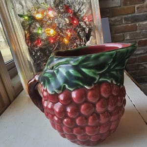 May include: A ceramic pitcher with a red, textured body resembling a raspberry. The rim is red, and the top is green with leaf detailing. The handle is brown. The pitcher is on a white surface.