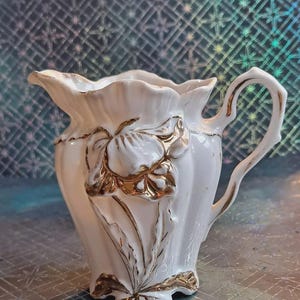 May include: White ceramic creamer with a gold floral design. The creamer has a handle and a ruffled top. The floral design is embossed and features a flower and leaves. The creamer is approximately 5 inches tall.