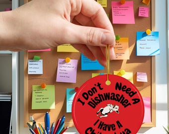 May include: A round, red ornament with the text "I Don't Need A Dishwasher I Have A Chihuahua" in black. A cartoon chihuahua is in the center. The ornament is held by a hand in front of a corkboard with sticky notes.