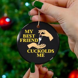 May include: Black round ornament with gold text that reads "MY BEST FRIEND CUCKOLDS ME". The ornament features illustrations of hands and keys. A gold string is attached for hanging.