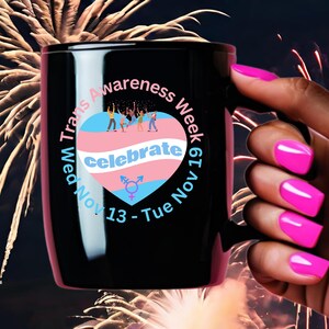 May include: Black ceramic mug with a white circle design. The circle features a pink, blue, and white heart with the text "Trans Awareness Week Nov 13 - Tue Nov 19" and "celebrate" inside the heart.  There are small figures of people in the heart.