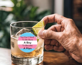 May include: A clear glass tumbler with ice and a round sticker featuring the transgender flag colors and the text "You Don't Need To Be Born A Boy To Become A Man." A hand holds a lime wedge over the glass.