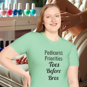May include: A light green t-shirt with the text "Pedicurist Priorities: Toes Before Bros" in black. The image also shows a woman smiling, nail polish bottles, and a manicure in progress.