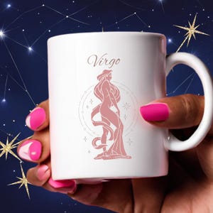 May include: White ceramic mug featuring the word "Virgo" in a script font and a pink illustration of a woman. The mug has a white handle and is held by a person with pink nail polish. The background has a starry night theme.