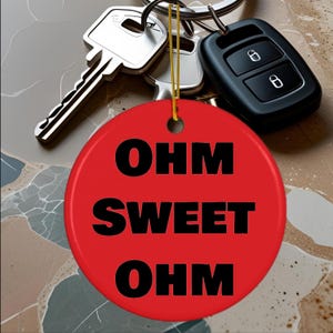 May include: A red, circular keyring tag with the words "OHM SWEET OHM" in black. The tag is suspended by a gold string next to keys and a black car key fob.