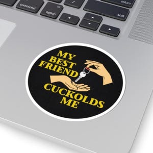 May include: A round sticker with the text "MY BEST FRIEND CUCKOLDS ME" in yellow, with a graphic of hands exchanging keys. The background is black with a white border. The sticker is on a laptop.