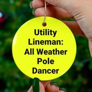 May include: A round, bright yellow ceramic ornament featuring the black text Utility Lineman: All Weather Pole Dancer. The ornament has a small hole at the top with a gold string attached for hanging, providing a smooth, glossy finish on the surface.