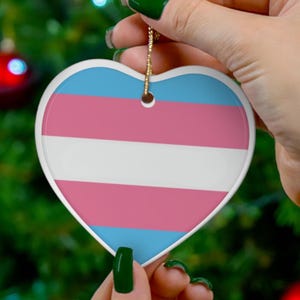 May include: Heart-shaped ceramic ornament featuring the transgender pride flag design. The ornament has a white border and a gold string for hanging. The flag design includes horizontal stripes of light blue, pink, and white.