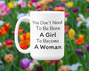 May include: White ceramic mug with a curved handle. The mug features the text "You Don't Need To Be Born A Girl To Become A Woman" in black, centered on the front. The background is a blurred field of colourful flowers.