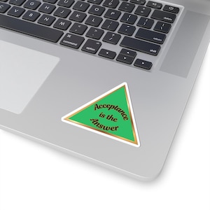 May include: A green triangle sticker with gold trim and the words "Acceptance is the Answer" in black script. The sticker is placed on a laptop.