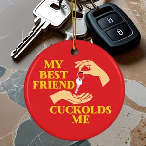 May include: A red, round keychain ornament with the text "MY BEST FRIEND CUCKOLDS ME" in yellow. The ornament features a graphic of hands exchanging a key. Other keys and a car key fob are visible in the background.