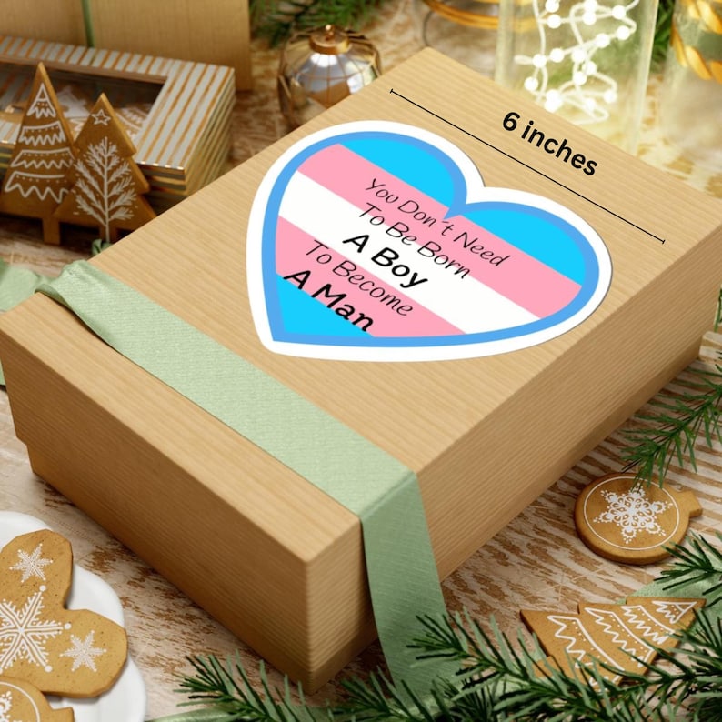 May include: A rectangular gift box with a light brown finish, wrapped with a light green ribbon. A heart-shaped sticker with the text "You Don't Need To Be Born A Boy To Become A Man" is on the box. The sticker is approximately 15 cm wide.