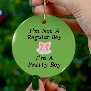 以下が含まれることがあります： 「I'm Not A Regular Boy, I'm A Pretty Boy」のテキストが黒で書かれた、丸い緑色のセラミックオーナメント。ピンクと青のリボンのグラフィックが中央に配置されています。吊り下げ用の金色の紐が付いています。