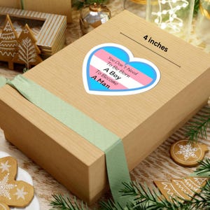 May include: A heart-shaped sticker, 10 cm in diameter, with the text "You Don't Need To Be Born A Boy To Become A Man" in a blue, pink, and white design. The sticker is on a brown gift box with a green ribbon.