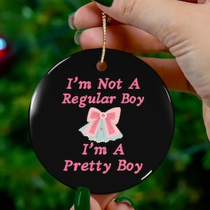 以下が含まれることがあります： 黒い丸いオーナメント。ピンク色の文字で「I'm Not A Regular Boy, I'm A Pretty Boy」と書かれています。ピンクと青のリボンのグラフィックが中央に配置されています。吊り下げ用の金色の紐が付いています。