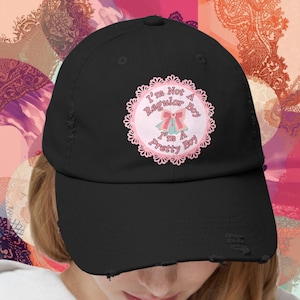 May include: Black baseball cap with a distressed finish. The cap has a circular patch with the text "I'm Not A Regular Boy I'm A Pretty Boy" in pink and white, with a bow graphic. The cap has a curved peak.