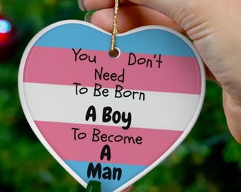 May include: Heart-shaped ornament with a transgender pride flag design. The ornament features the colours pink, white, and blue, with the text "You Don't Need To Be Born A Boy To Become A Man" in black lettering. A gold string is attached for hanging.